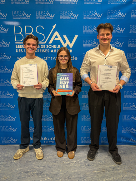 Due ragazzi e una ragazza sono in posa con i loro diplomi davanti a una parete blu con il logo della scuola professionale di Ahrweiler
